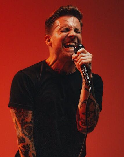 Tattooed male performer sings energetically into a microphone against a bold red background.