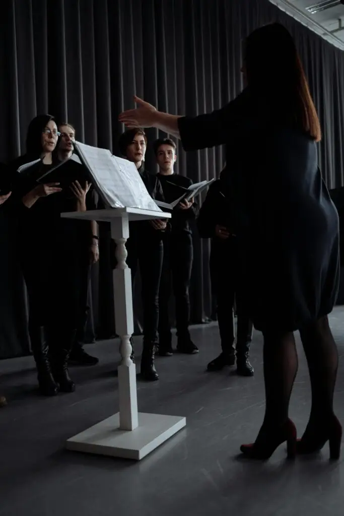 A choir rehearses with a conductor standing by a music stand indoors, all in black attire.