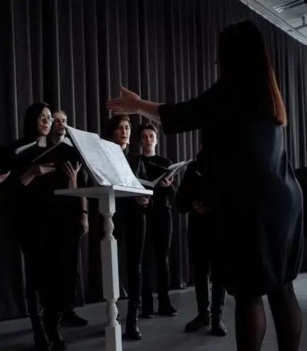 A choir rehearses with a conductor standing by a music stand indoors, all in black attire.